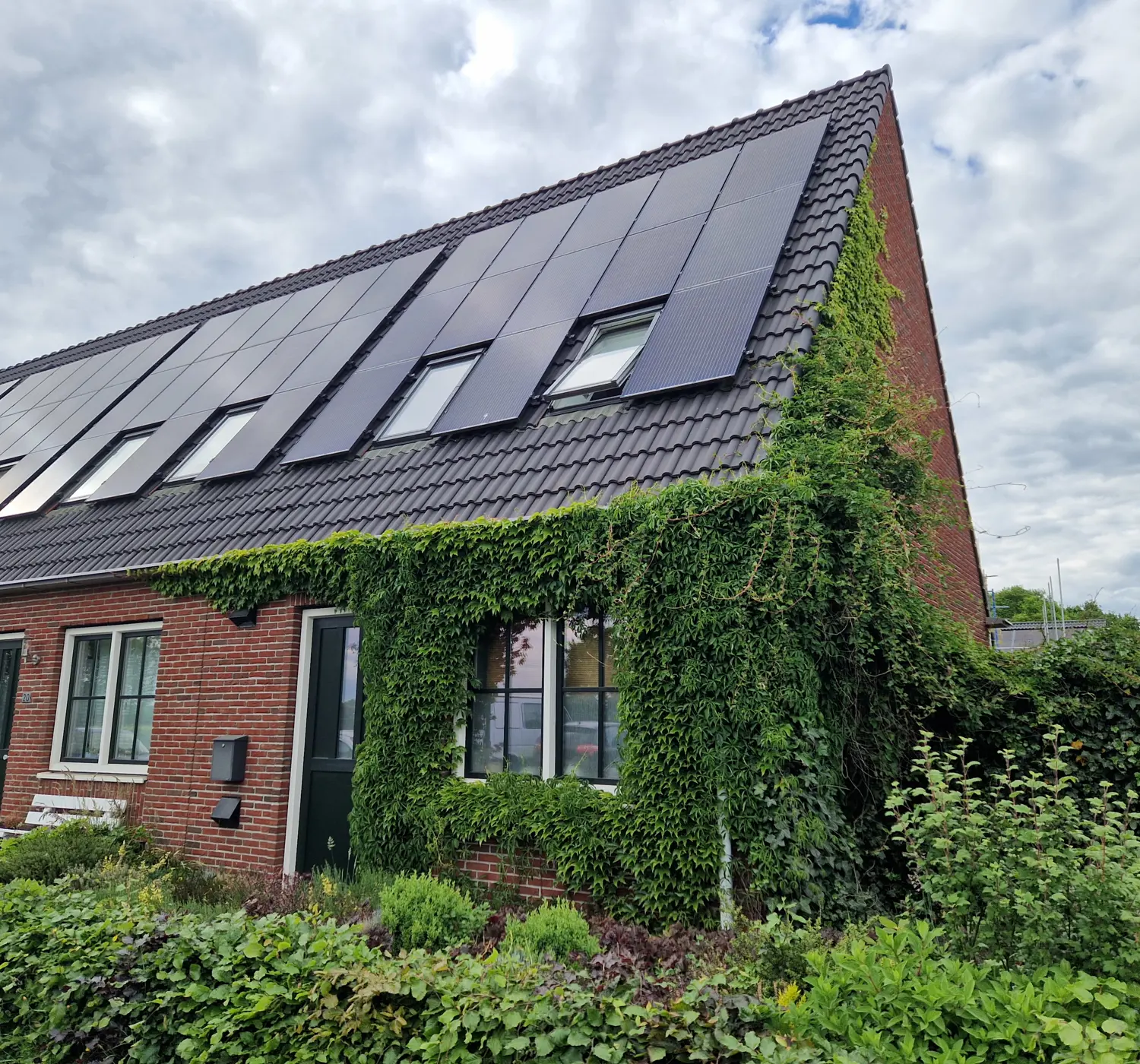 <img src="woning.jpg" alt="Moderne rijtjeswoning met schuin dak voorzien van zonnepanelen. De gevel is deels bedekt met groene klimplanten. Voor de woning ligt een verzorgde tuin met struiken en planten. De scène is verlicht door diffuus daglicht onder een bewolkte hemel.">