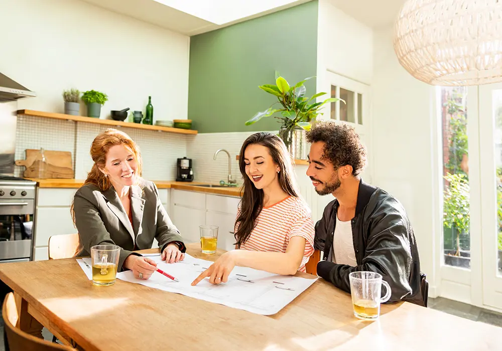 Adviseur zit met klanten aan de keukentafel. De adviseur wijst iets aan op een document.