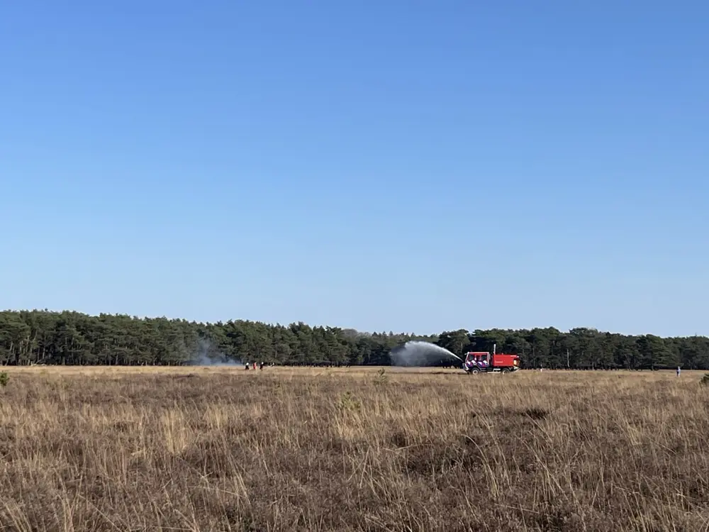 <img src="brandbestrijding.jpg" alt="Droog grasveld met op de achtergrond een bos. Rechts in beeld staat een rode brandweerwagen die water spuit op een plek waar rook zichtbaar is. Enkele mensen, vermoedelijk brandweerlieden, staan bij de rook. De lucht is helder en blauw.">